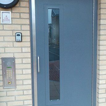 SINGLE COMMUNAL ENTRANCE STEEL DOOR FABRICATED TO LPS1175 SR2 OR PAS24 STANDARD Secure by Design accredited grey security door with a vertical window, intercom panel, and access control system on a brick wall.