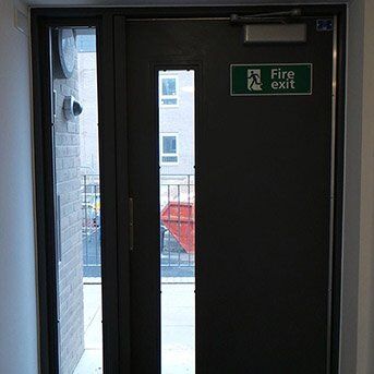 FIRE EXIT STEEL DOOR FABRICATED AND INSTALLED BY PREMIER SECURITY CONSULTANTS OF ILFORD Fire exit door with a clear view of the outside, featuring a "Fire exit" sign, part of a Secure by Design accredited installation for enhanced safety in residential and commercial properties.