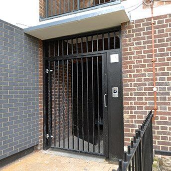 PUBLIC ACCESS GATE FABRICATED IN STEEL Secure by Design accredited black security gate with vertical bars and access control panel, set in a brick wall environment, showcasing high-standard safety features for residential and commercial properties.