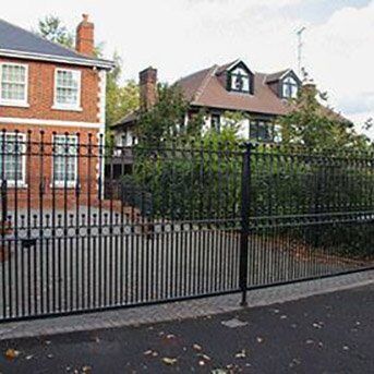 STYLISH HIGH SECURITY STEEL GATES FOR HOME PROTECTION Black security gate with vertical bars, providing access control to residential properties, surrounded by greenery and traditional brick houses, illustrating Secure by Design accredited security solutions.