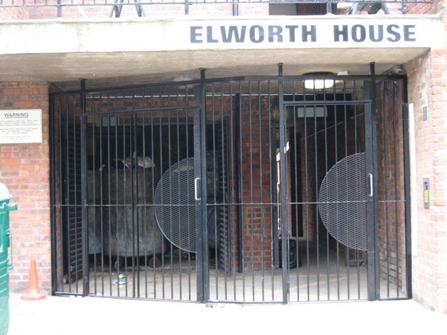 Black security gates at Elworth House entrance, featuring circular mesh designs, providing access to a bin store area, emphasizing high-security design in communal settings.
