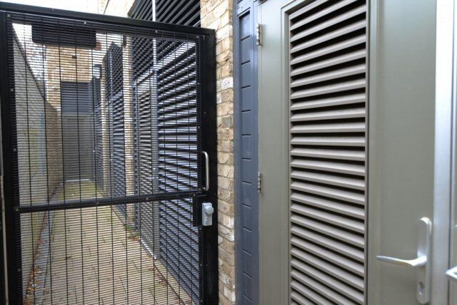 Secure by Design accredited security gate with a lock, adjacent to louvered doors in a brick-lined corridor, emphasizing durability and safety features.