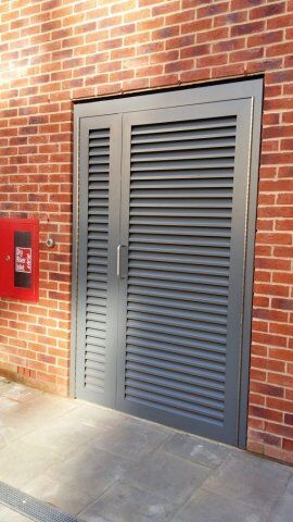 Gray louvre security door installed in a brick wall, adjacent to a red mailbox, showcasing Secure by Design accredited features for enhanced safety in residential or commercial properties.