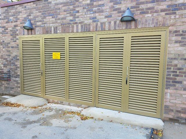 Secure by Design accredited louvre doors in a muted gold finish, installed on a brick wall, featuring a warning sign and dual handles, illustrating robust security solutions for communal areas.