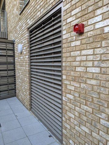 Louvre security door installed on a brick wall, featuring horizontal slats for ventilation and a red alarm light nearby, highlighting secure access solutions for residential and commercial properties.