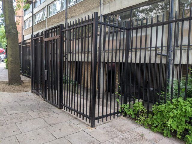 Black security gate with vertical bars, providing access to a building's entrance, surrounded by greenery and concrete paving, emphasizing safety and design compatibility in urban settings.