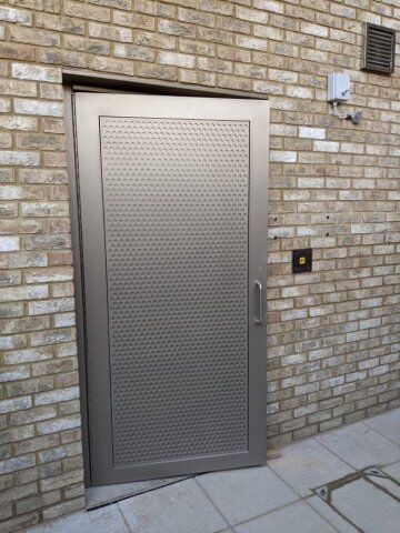 Industrial steel security door with textured surface, installed in a brick wall, featuring an access control panel nearby, emphasizing high-security design standards.