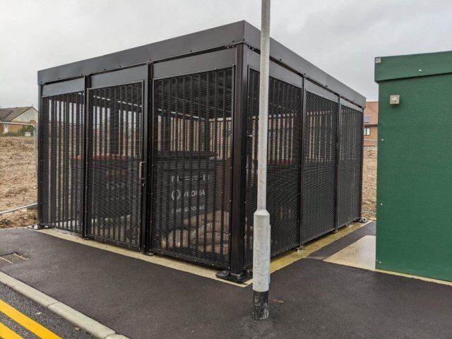 Secure by Design accredited bin store enclosure with metal mesh walls and a roof, positioned beside a green utility box on a paved area.