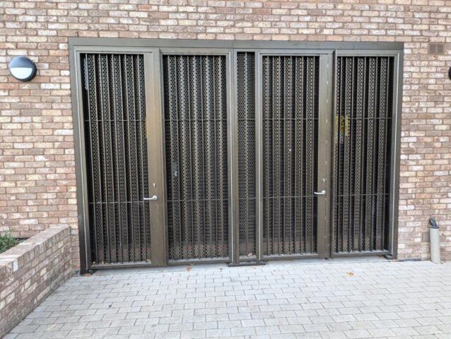 Steel security doors with mesh design, featuring multiple access points, installed in a brick wall setting, highlighting Secure by Design accreditation for enhanced safety.