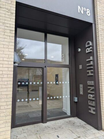 Secure by Design accredited communal steel entrance featuring glass doors, numbered "N°8," with the street name "HERNE HILL RD" prominently displayed, showcasing security and modern design elements.
