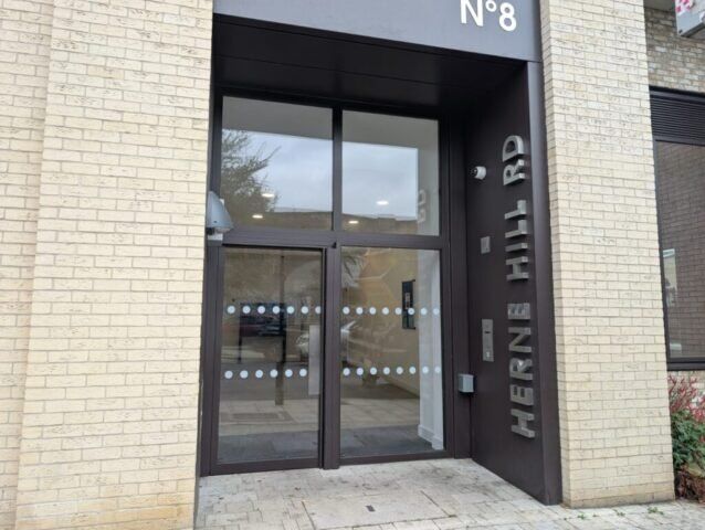 Entrance of a building featuring glass doors with security features, displaying the address "HERNE HILL RD" and numbered "N°8," emphasizing modern design and accessibility.