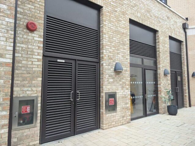 Secure by Design accredited communal steel doors with louvre design, brick wall background, and adjacent fire safety equipment, showcasing high-standard security solutions for residential and commercial properties.
