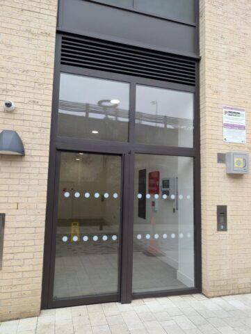 Secure by Design accredited communal steel doors with glass panels and circular frosted windows, set in a modern building entrance with intercom and signage.