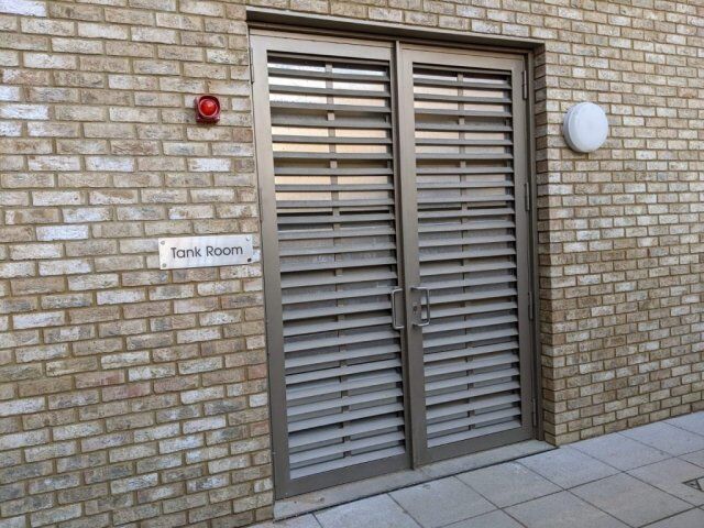 Louvre security doors with a "Tank Room" sign, featuring a brick wall background and a red alarm light, showcasing durable design for enhanced safety in building installations.