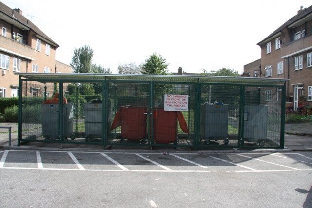 Secure bin store with multiple waste containers and a sign stating "No parking in front of this store on Thursdays," surrounded by residential buildings and greenery.