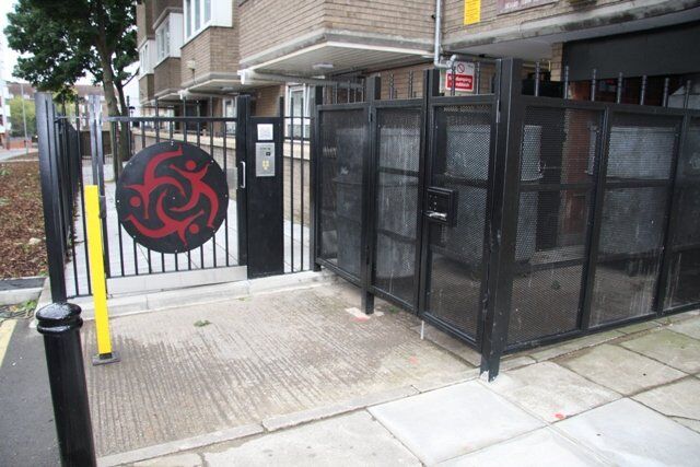 Security gate with red emblem, featuring mesh panels and access control system, situated in a residential area, showcasing Secure by Design principles for enhanced safety.