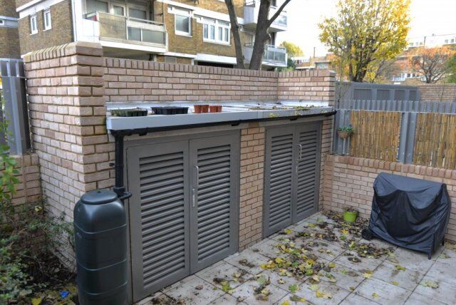 Secure by Design accredited bin store with louvered doors, integrated countertop, and adjacent water tank, set in a residential area.