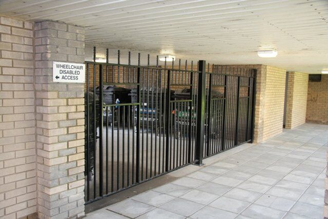 Wheelchair accessible gate in a secure bin store area, featuring black metal bars and a sign indicating disabled access.