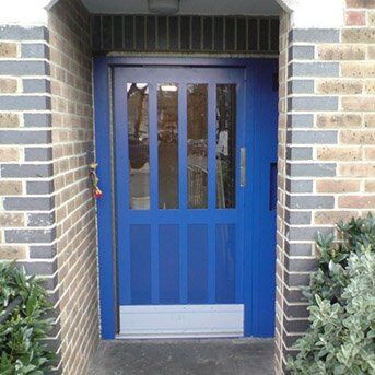 Secure by Design accredited blue security door with vertical glass panels, framed by brick wall and greenery, showcasing high-standard security features.