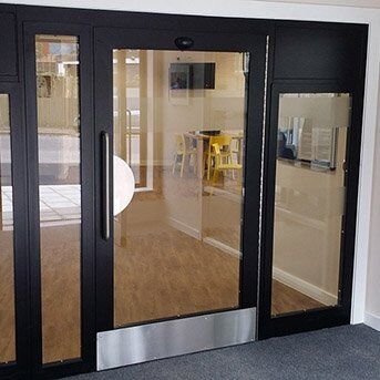 Secure by Design accredited glass and steel communal doors with a sleek black frame, featuring a large circular handle and transparent panels, showcasing a modern interior space.