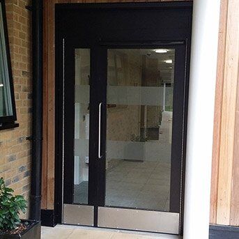 Secure by Design accredited black steel communal door with glass panels and a polished bottom section, showcasing modern security features in a residential setting.