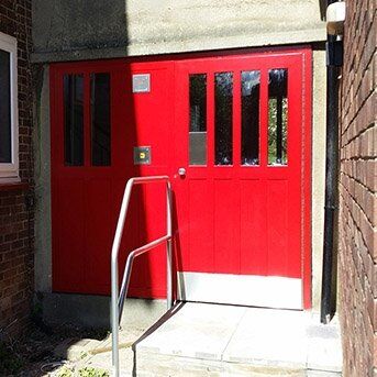 Red security door with glass panels and access control system, surrounded by a concrete wall and steps, representing Secure by Design accredited installations for enhanced safety.