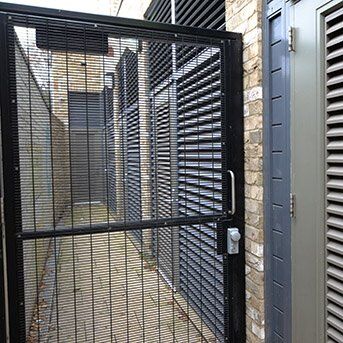 Secure by Design accredited black security gate with mesh design, leading to a narrow pathway between brick walls, showcasing a practical application for enhanced safety in residential or commercial properties.