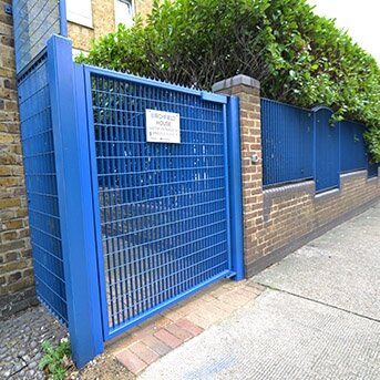 Blue security gate with mesh design, leading to a property, featuring a sign that reads "SECURED HOUSE," surrounded by greenery and brick wall, showcasing a Secure by Design accredited installation.