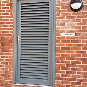 Louvre security door in grey, installed in a brick wall, labeled 'Gas Meters', showcasing Secure by Design features for enhanced safety and access control.
