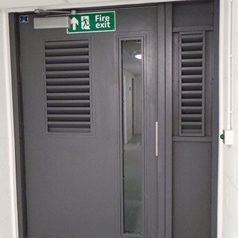 Secure by Design accredited fire exit door with ventilation louvres and clear viewing panel, emphasizing safety and compliance in security installations.