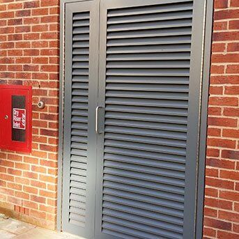 Louvre security door in grey against a brick wall, featuring a handle and adjacent red fire alarm box, exemplifying Secure by Design accredited products for enhanced building safety.