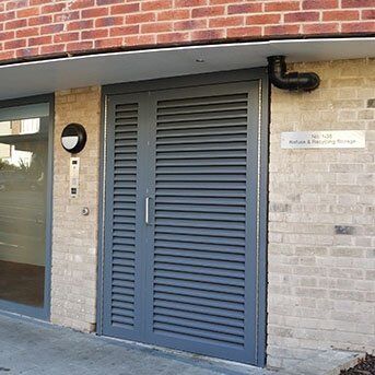 Secure by Design accredited louvre security door in grey, installed in a brick building, featuring intercom and signage for secure access.