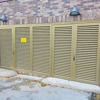 Louvre security doors in a beige finish, featuring horizontal slats and a warning sign, positioned against a brick wall, highlighting secure access solutions for communal areas.