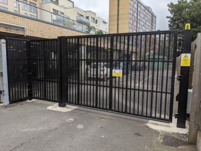 Automated security vehicle gates with black mesh design, featuring warning signs, providing access control to a parking area surrounded by residential buildings.