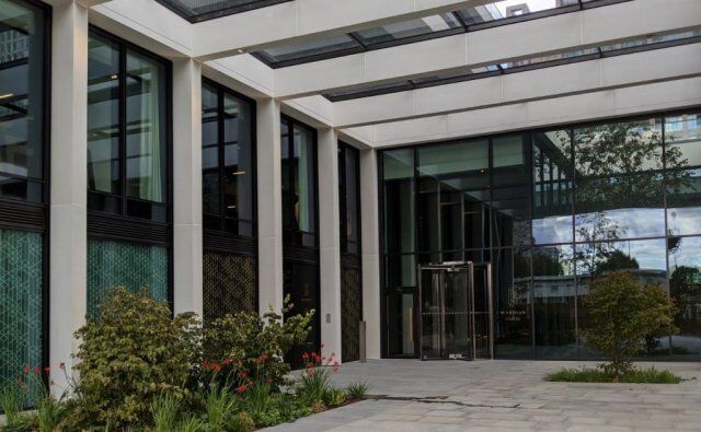 Modern building entrance featuring secure glass curtain walling, landscaped greenery, and a revolving door, emphasizing security and design aesthetics in urban architecture.